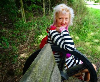 Portrait of smiling mature woman sitting by metallic wheel in forest