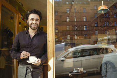 Portrait of happy barista holding coffee cup while standing at doorway