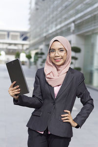 Portrait of confident businesswoman holding digital tablet while standing on street outside office building