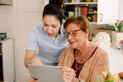 Retired elderly woman looking while young female volunteer using digital tablet in nursing home kitchen