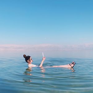 Woman reading paper while floating in sea against blue sky