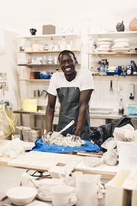 Portrait of happy young artist wearing apron standing with clay on table in art class