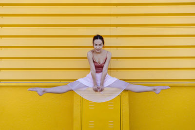 Young woman doing splits while ballet dancing on seat against yellow wall