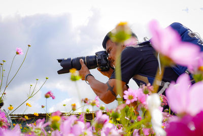 Side view of man photographing by flowering plant against sky