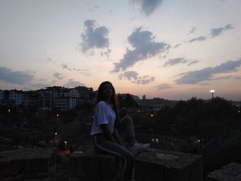 Woman sitting on cityscape against sky during sunset