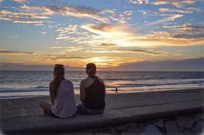 Rear view of female friends sitting on retaining wall at beach against sky during sunset