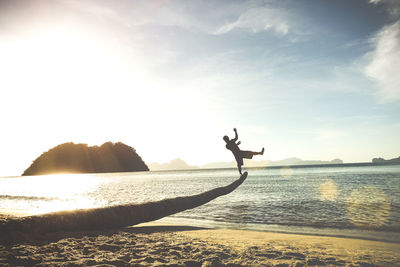 Silhouette man balancing on log at sea shore against sky