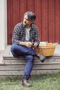 Thoughtful farmer with wicker basket holding mobile phone while sitting on porch at yard