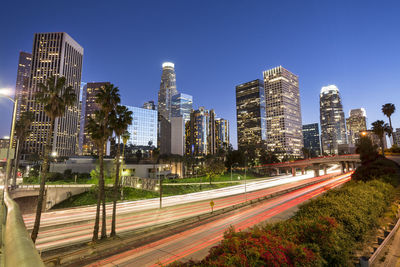 Light trails on road by illuminated buildings against clear sky