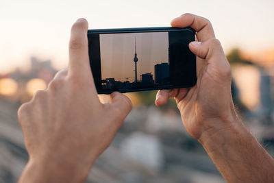 Cropped hands of man photographing tower with mobile phone in city during sunset