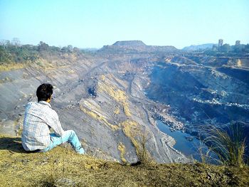 Rear view of man sitting on cliff against open-pit mine