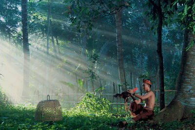 Side view of man sitting with rooster in forest