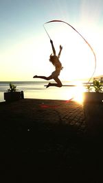 Full length of woman jumping while waving ribbon against sea during sunset