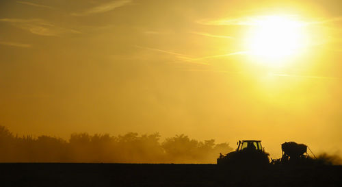 Silhouette of a tractor sowing seeds in a field in a cloud of dust against the background.