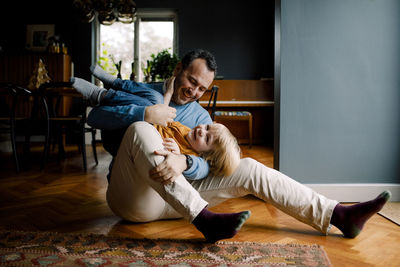 Full length of happy playful father carrying daughter while sitting on hardwood floor at home
