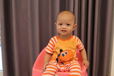 Portrait of cute baby boy sitting on seat against curtain at home