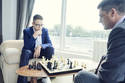 Dad and son are sitting at a table by a wide window and playing chess making moves with pieces