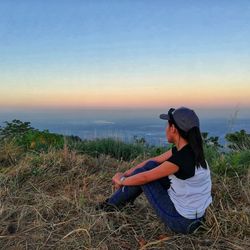 Young woman sitting on grass against sky during sunset