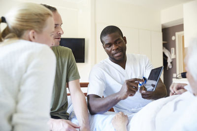 Male nurse discussing over digital tablet with couple while senior man reclining on hospital bed