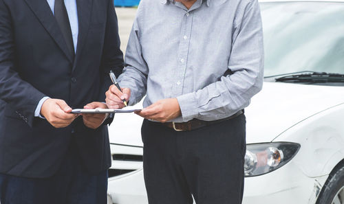 Midsection of man signing on paper held by businessman against car