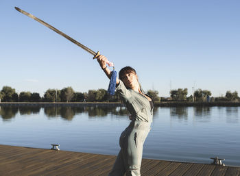 Young woman doing martial arts with sword in park by lake on sunny day