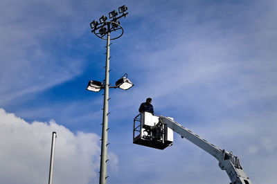 Low angle view of worker in cherry picker by street light against sky