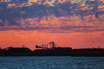 Silhouette cranes at harbor against sky during sunset