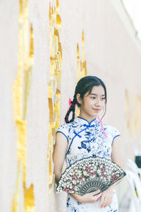 Teenager girl holding hand fan standing against wall