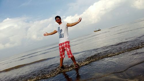 Tilt image of young man standing with arms outstretched on sea shore at beach against cloudy sky