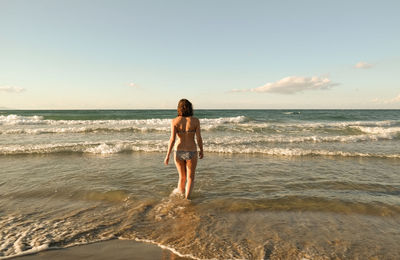 Rear view of young woman in bikini walking at beach against sky