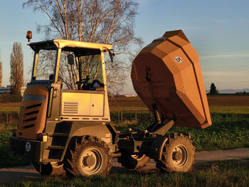 A  dump truck with its trough raised stands on a dirt road outside the city in the evening light. 
