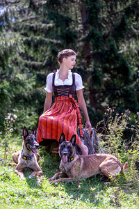 Full length of young woman with dogs sitting against trees in forest