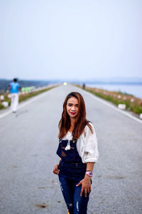 Portrait of smiling young woman standing on road against sky