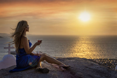 Woman sitting on beach by sea against sky during sunset