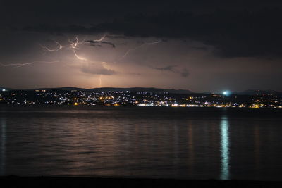 Illuminated city by sea against sky at night