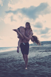 Portrait of shirtless man carrying girlfriend on shoulder at beach against sky during sunset