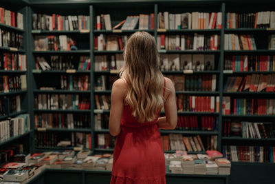 Back view of anonymous elegant woman in sundress standing against table with assorted textbooks in bookstore