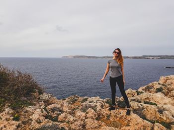 Portrait of woman wearing sunglasses standing on rock by sea against sky