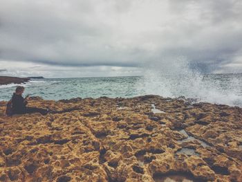 Side view of woman siting on rocky shore against cloudy sky