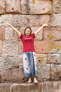Portrait of woman with arms raised standing against stone wall