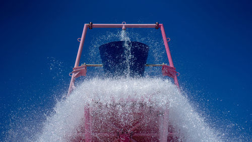 Low angle view of bucket pouring water against clear blue sky