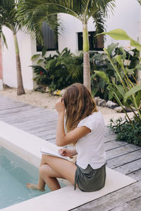 Rear view of woman sitting on poolside writing on a notebook 