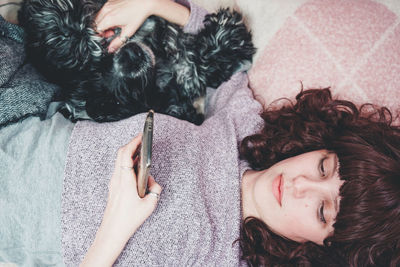 Woman using phone while lying with dog on bed at home