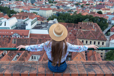 A girl with long hair in a hat sits on the edge of a tall building and looks at the city of graz. 