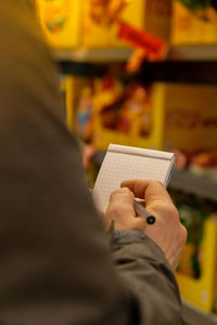 Female hands holding paper notebook of shopping list, blurred shelves on background. partial view of