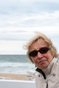 Portrait of senior woman wearing sunglasses standing at beach against cloudy sky