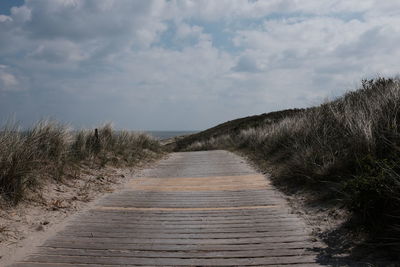 Empty boardwalk amidst grass at beach against sky