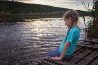 Girl sits on pontoon bridge at lake and enjoys warm sunny evening, happy summertime, countryside