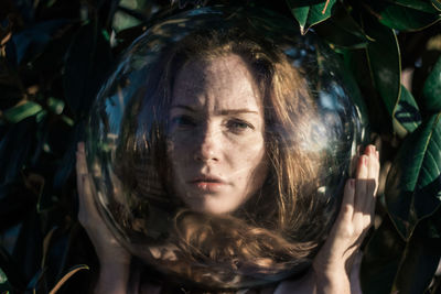 Close-up portrait of young woman wearing glass ball on head against leaves