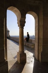 Woman sitting on retaining wall seen through arch against sky